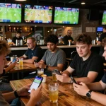 Pengguna SBOBET memasang taruhan di suasana bar olahraga yang ramai, berinteraksi dengan aplikasi permainan seluler.